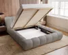 A modern bedroom with a grey upholstered bed frame, its storage compartment open, revealing the slatted base and mattress space. The bed is placed on a patterned rug in front of a slatted wooden wall and a window. Náhled obrázku produktu