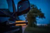 A close-up shot of a car's side mirror and a glowing orange LED light attached to the car's body, with a blurred background of trees and a road at dusk. Produktbild-Vorschau