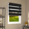 A window with black and white zebra blinds, showing a green lawn and a wooden swing set outside. A shelf with framed pictures and bowls is to the left, and a desk with a vase of sunflowers is to the right. Voir les images du produit