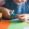 A child's hands are shown using a pair of training scissors to cut a piece of orange paper. The scissors are blue and green. There is green and orange paper on a table in front of the child. Voir les images du produit