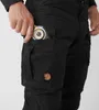 A person's hand is shown placing a compass into the pocket of black outdoor trousers. Produktbild-Vorschau