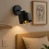 A modern black wall lamp is mounted on a wall above a wooden headboard and pillow in a bedroom setting. Produktbild-Vorschau