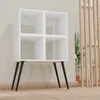 A white cube shelf unit with four compartments, supported by four black metal legs, stands on a wooden floor against a light-colored wall. A slatted wooden screen and a potted plant are visible in the background. Produktbild-Vorschau