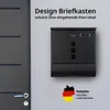 A black mailbox mounted on a light gray wall, next to a dark gray door. Produktbild-Vorschau