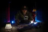 A person in tactical gear is looking at a smartphone in a dimly lit room, with firearms and ammunition scattered on the surface in front of them. Produktbild-Vorschau