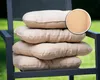 A stack of four round beige seat cushions on a dark gray chair, with a blurred green background. Produktbild-Vorschau