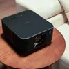 A black projector sits on a wooden table next to a couch. Anteprima dell’immagine prodotto