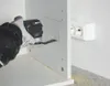 A person is using a power tool to cut a hole in a white panel, with a power strip visible on the wall in the background. Podgląd zdjęcia produktu