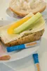 A close-up of a sandwich with cheese, cucumber, and what appears to be bell pepper, served on a plate with two salt packets. Anteprima dell’immagine prodotto