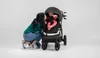 A woman is kneeling next to a baby stroller, leaning in to kiss the baby inside. The stroller is black with a brown handle. Anteprima dell’immagine prodotto