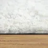 A close-up, side view of a fluffy white rug on a wooden floor. Produktbild-Vorschau