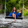 A woman is sitting on a swing over a river, wearing a blue dress. Náhled obrázku produktu