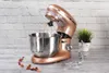 A rose gold stand mixer with a whisk attachment sits on a table in front of a white brick wall. A small potted plant is to the right of the mixer. Anteprima dell’immagine prodotto