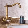 A close-up shot of a vintage-style brass faucet with two handles, set against a marble-patterned background. A small gray dispenser and folded towels are visible to the right. Anteprima dell’immagine prodotto