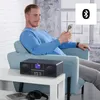 A man is sitting in a chair, looking at his phone, with a radio on a table in front of him. Produktbild-Vorschau