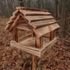 A wooden bird feeder stands on a wooden pole in a forest setting with fallen leaves on the ground. Podgląd zdjęcia produktu