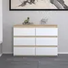 A modern dresser with six drawers, featuring a white front and a wood-grain frame, is displayed in a room with textured wallpaper and a dark tiled floor. Produktbild-Vorschau