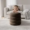 A young girl is resting her arms on a stack of brown ottomans in a living room. Produktbild-Vorschau
