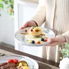 A person is holding a plate with a dessert and fruit, with another plate of food in the foreground. Produktbild-Vorschau
