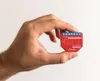 A hand holds a small red electronic device with blue connectors. Produktbild-Vorschau