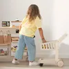 A young girl plays with a wooden toy shopping cart in front of a toy store counter. Produktbild-Vorschau
