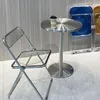 A clear, well-lit studio shot of a small round table with a coffee cup on it, and a transparent folding chair next to it. In the background, two other folding chairs are visible, one blue and one brown. Produktbild-Vorschau