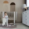 A baby playing with a blanket next to a crib in a nursery. Produktbild-Vorschau