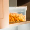 A clear plastic food storage container filled with corn flakes, placed on a shelf next to a peach-colored cabinet. Produktbild-Vorschau