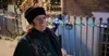 An elderly woman stands in the snow, wearing a hat and scarf, with Christmas lights in the background. Náhľad obrázku produktu