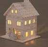 A white wooden house with illuminated windows and a small decorative tree and figure in front. Podgląd zdjęcia produktu