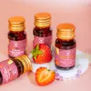 Three small bottles of liquid collagen with strawberries and a beaded bracelet on a pink background. Produktbild-Vorschau