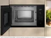 An open built-in microwave oven is shown in a kitchen setting, with a partial view of a countertop with plants and nuts to the right. Náhľad obrázku produktu