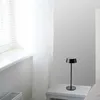 A black table lamp sits on a white nightstand next to a bed. A window with blinds is visible on the left. Náhľad obrázku produktu