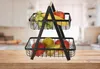 A two-tiered fruit basket filled with various fruits, placed on a wooden table in a kitchen setting. Anteprima dell’immagine prodotto