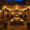 An outdoor patio area at dusk, set up for a party with a table and chairs illuminated by string lights. Produktbild-Vorschau