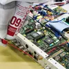 A gloved hand is holding a can of KONTAKT CHEMIE Kontakt 60 spray and spraying it onto a complex electronic circuit board. Produktbild-Vorschau
