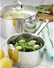A close-up view of two stainless steel pots on a wooden table, one containing green beans and olives, the other with a lid on and steam rising. Náhľad obrázku produktu
