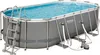 An oval-shaped above-ground swimming pool filled with blue water, set against a white background. The pool has a gray frame and a ladder on the left side. A filter pump and hoses are visible at the bottom right. Anteprima dell’immagine prodotto