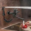 A black kitchen faucet with a sprayer is shown mounted on a wall above a sink. Water is spraying from the faucet and the sprayer. A person is washing strawberries in the sink. Náhled obrázku produktu