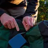 A person's hands are shown interacting with a small blue pouch attached to a green and black garment. Produktbild-Vorschau