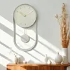 A minimalist pendulum wall clock with a light pastel green face and gold hands is displayed on a white wall, casting shadows from sunlight. Decorative vases and dried flowers are arranged on a wooden cabinet below. Voir les images du produit