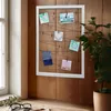 A white frame with strings and clips holding photos and cards, placed on a wooden wall next to a window and a potted plant. Náhled obrázku produktu