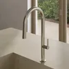 A modern kitchen sink faucet with a curved spout and a single lever handle, set against a neutral countertop and a blurred background of a window with greenery. Produktbild-Vorschau