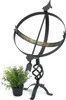 A black metal sundial with Roman numerals on a white background, next to a small potted plant. Anteprima dell’immagine prodotto