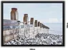 A white bicycle is leaning against a wooden sea defense structure on a pebble beach, with the sea and sky in the background. Náhled obrázku produktu