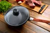 A frying pan with a glass lid and a wooden handle is placed on a wooden table, with raw meat and vegetables in the background. Voir les images du produit