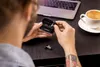 A person is holding a pair of wireless earbuds and their charging case, with a laptop and a cup of coffee on a desk. Produktbild-Vorschau