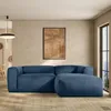 A modern living room with a blue corduroy sectional sofa and ottoman, a framed abstract artwork on the wall, and a large window with a view. Produktbild-Vorschau
