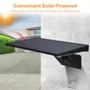 A solar-powered wall light mounted on a textured wall, with a house and driveway in the background. Produktbild-Vorschau