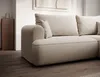 A close-up view of a cream-colored sectional sofa with plush cushions, placed on a wooden floor in a well-lit room. A wooden cabinet is visible in the background. Produktbild-Vorschau